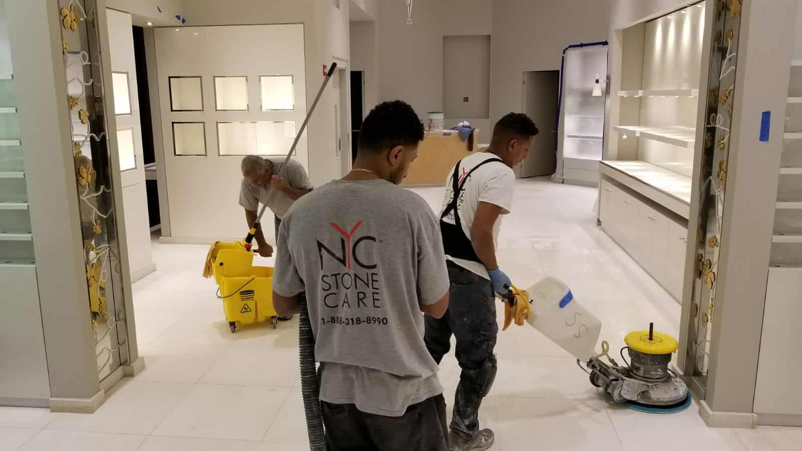 Three workers clean and polish a shiny white floor in a modern, well-lit room. One uses a floor polishing machine for expert stone restoration NYC style, while another mops and the third carries cleaning equipment. Shelves and display cases line the walls.