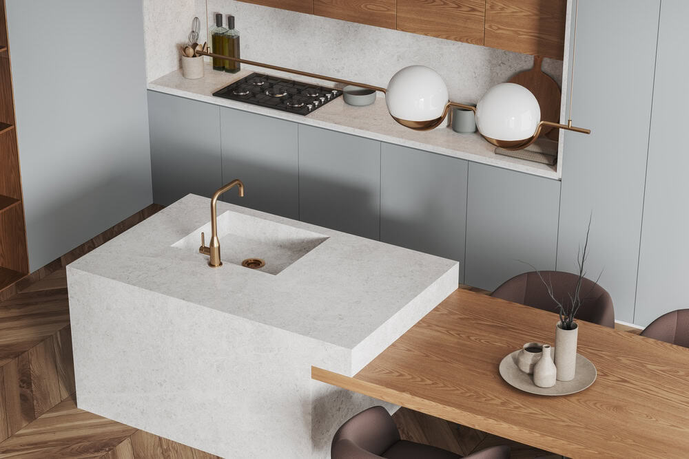 Modern kitchen with a marble island featuring a built-in sink and gold faucet, gray cabinets, a wooden dining table, and minimalist decor. Expert stone restoration NYC keeps the marble looking pristine alongside stylish round white lamps and potted plants.